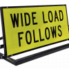 Pillot Vehicle Sign Rig » Highway 1 Bright yellow warning sign reading 'WIDE LOAD FOLLOWS' mounted on a black metal frame and stand.