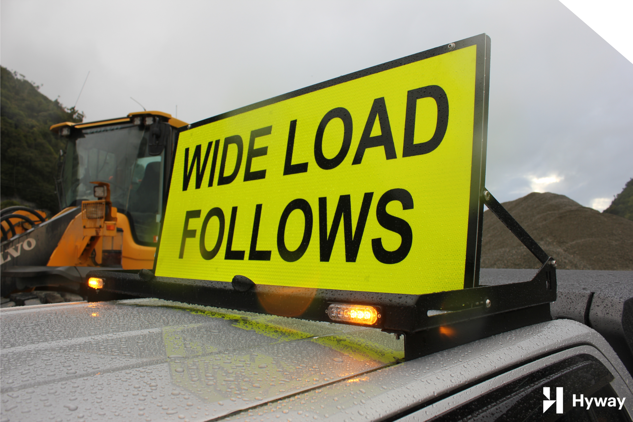 Sign Rig 2 » Highway 1 Bright yellow road sign reading 'WIDE LOAD' on a construction vehicle in a rainy setting with water droplets on the car hood.