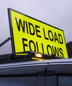 Sign Rig » Highway 1 Bright yellow highway sign reading 'WIDE LOAD FOLLOWING' mounted on a vehicle, cloudy sky in the background.