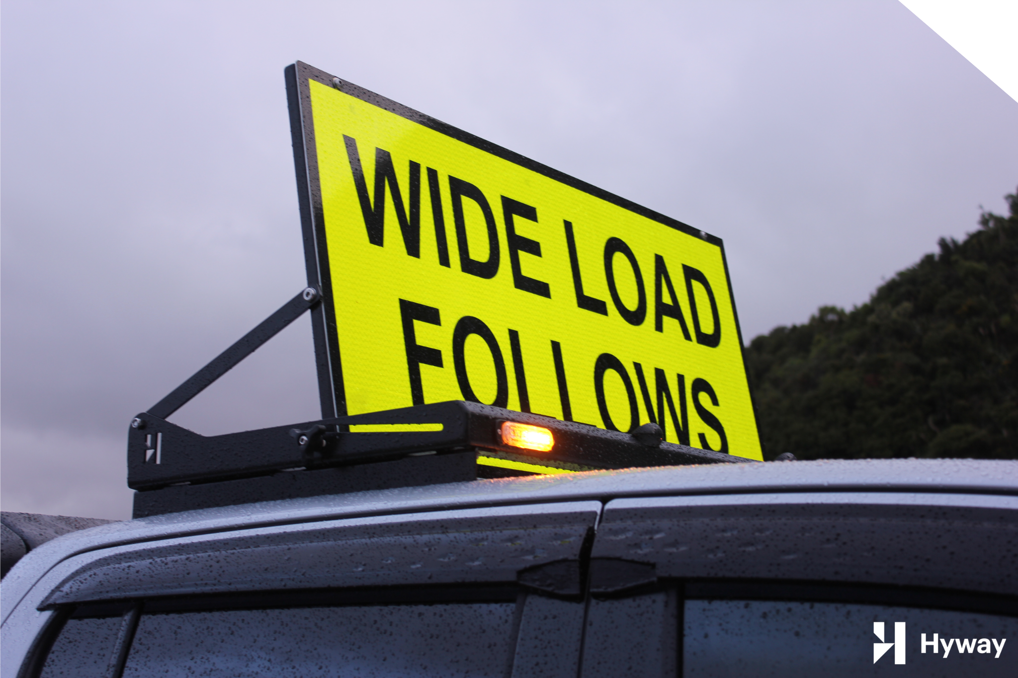 Sign Rig » Highway 1 Bright yellow highway sign reading 'WIDE LOAD FOLLOWING' mounted on a vehicle, cloudy sky in the background.